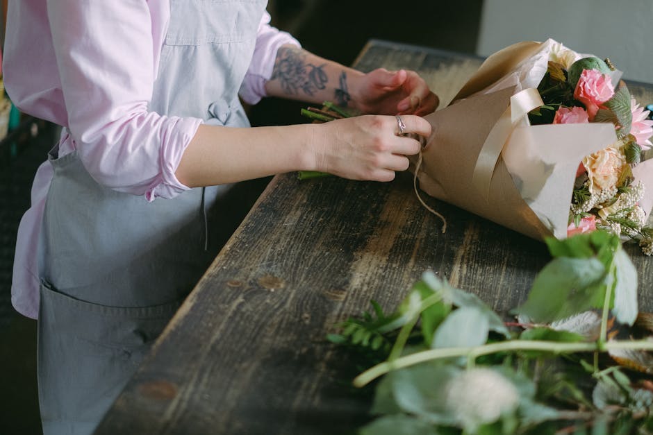 A person wearing a light pink long-sleeve shirt and a grey apron is arranging a flower bouquet on a rustic wooden table. The bouquet is wrapped in brown paper and tied with a cream ribbon, featuring p