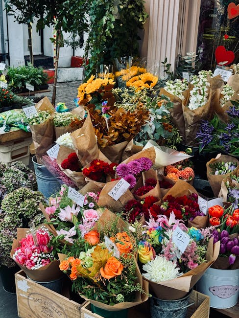 A vibrant display of freshly arranged flower bouquets situated outdoors near a garden setting. The bouquets feature a variety of flowers including bright yellow sunflowers, pink and red roses, purple 