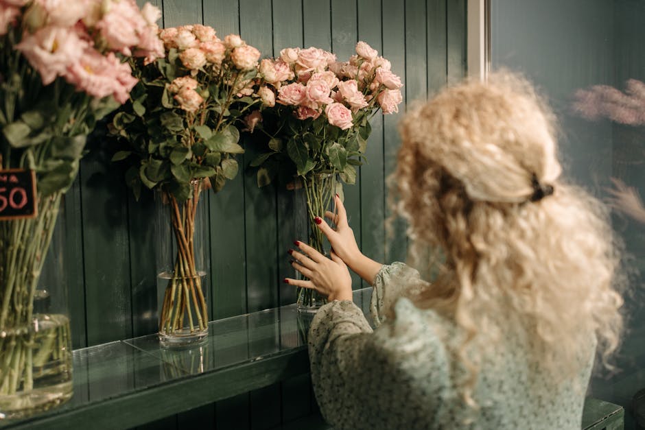 A woman with curly blonde hair, wearing a beige floral top, arranging a bouquet of pale pink roses in a tall glass vase inside a florist shop. The arrangement features fresh roses with slightly ruffle