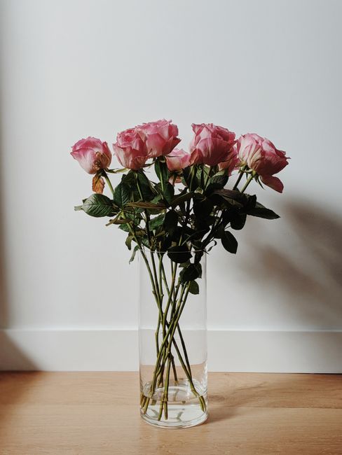 A clear glass vase filled with fresh pink roses featuring delicate, slightly ruffled petals and dark green leaves, arranged in a loose bouquet on a wooden surface against a plain white wall. The roses